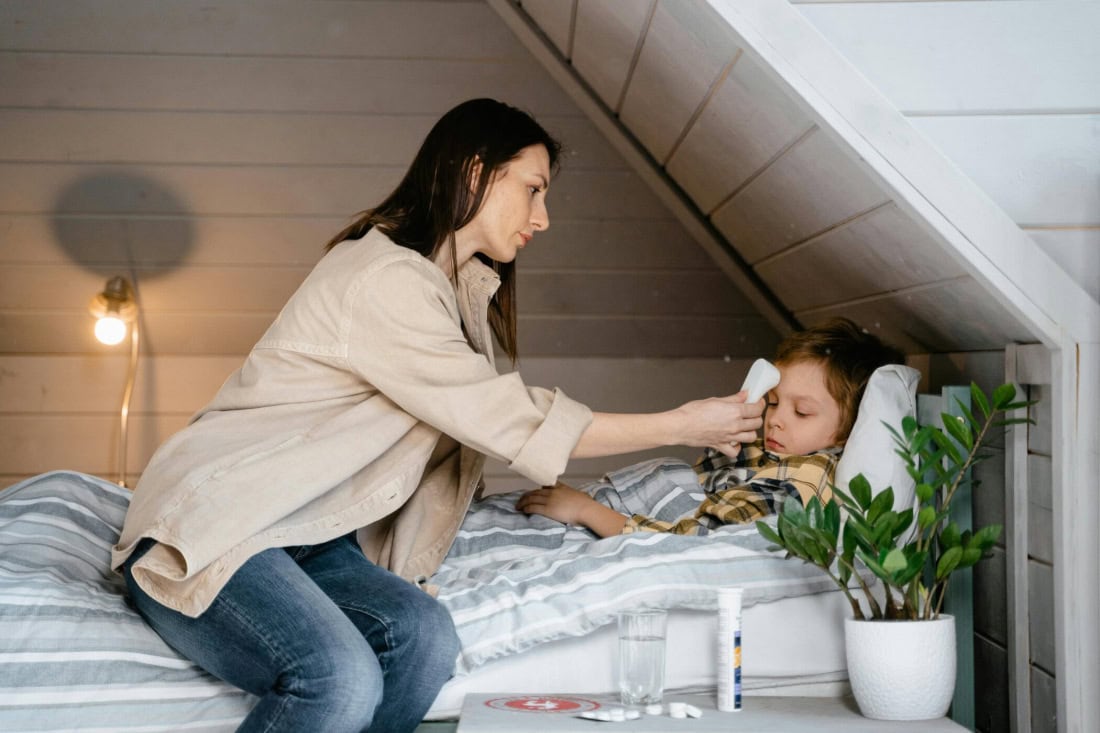 Koortsverzorging bij een kind in een slaapkamer met moeder, thermometer en medicatie.
